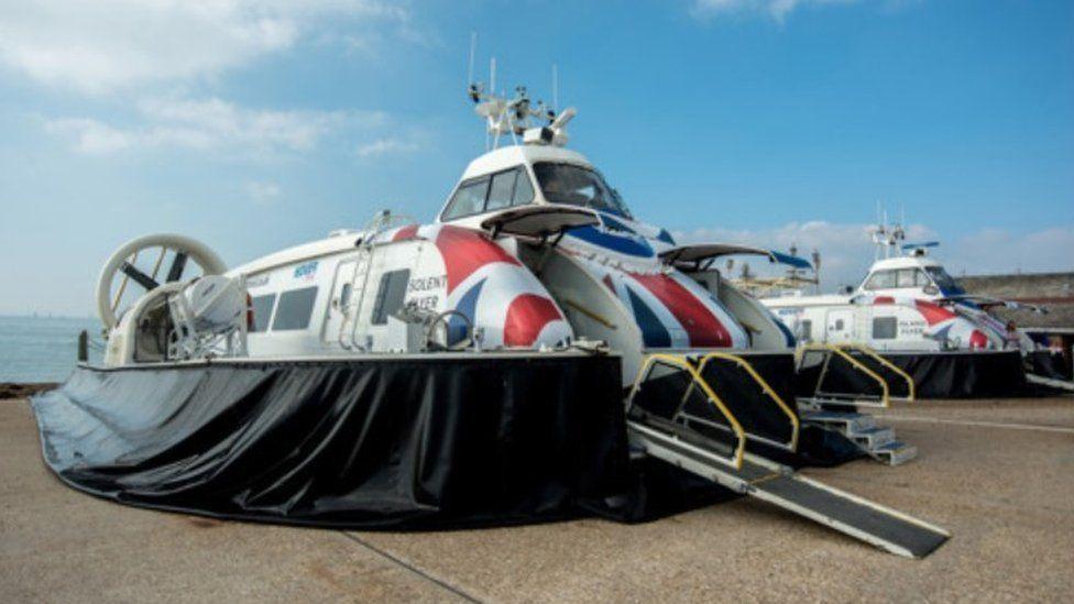 Two hovercrat on a concrete area - they have black skirts and ramps down - the top passenger areas of the craft are in red white and blue.