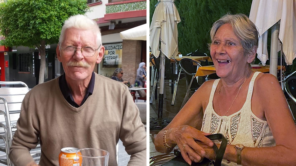 Terri (right) is wearing a white vest top and smiling with a bag on her lap. She has short, grey hair, or her hair is possibly tied back. Don is wearing glasses and has a moustache and is wearing a light brown jumper