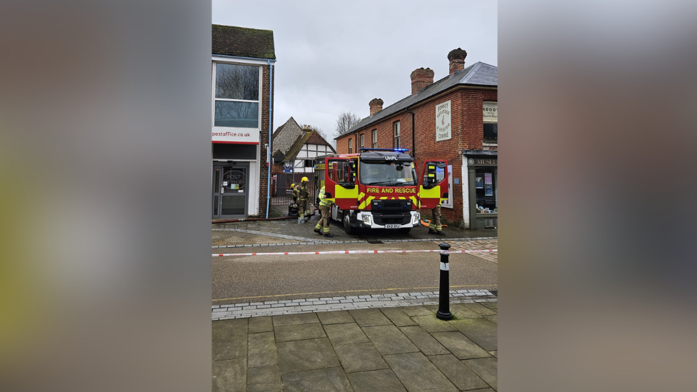 A fire engine is seen blocking a narrow street in between two small commercial buildings. Fire crews are seen standing around it and red and white tape is seen blocking the street in front. 