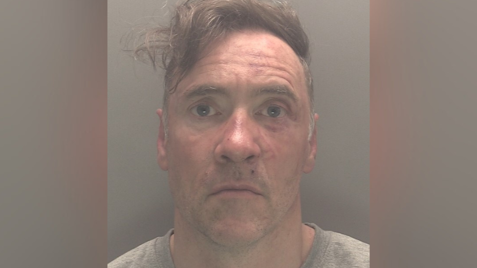 A mugshot of Paul Doyle wearing a grew jumper looking stunned. His hair, a long brown fringe, is dishevelled and to the side. He is wearing a grey t-shirt.