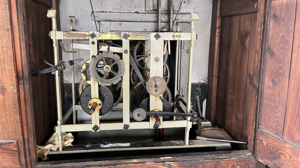 Image shows the inside workings of the one-handed clock in a wooden cabinet with lots of big and small cogs