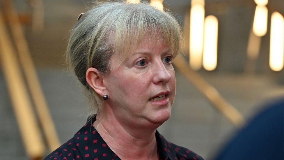 Shona Robison, who has blonde hair tied back, speaks at the foot of a set of stone stairs. She is wearing a dark top with red polka dots and is visible from the shoulders up. 