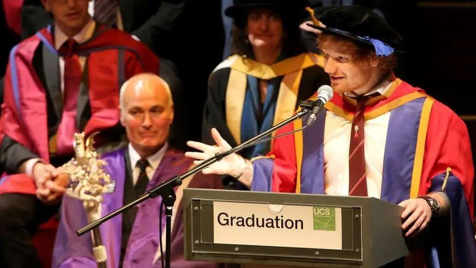 ed Sheeran in a cap and gown speaking at a podium.