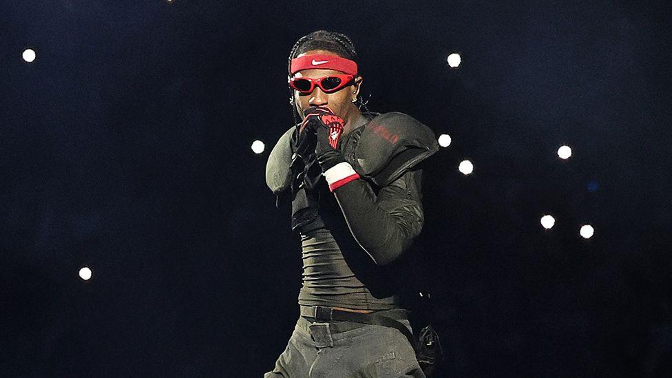 Travis Scott, a man performing on stage, wearing all black, with red framed sunglasses and a Nike bandana on his forehead, with spotlights in the background.