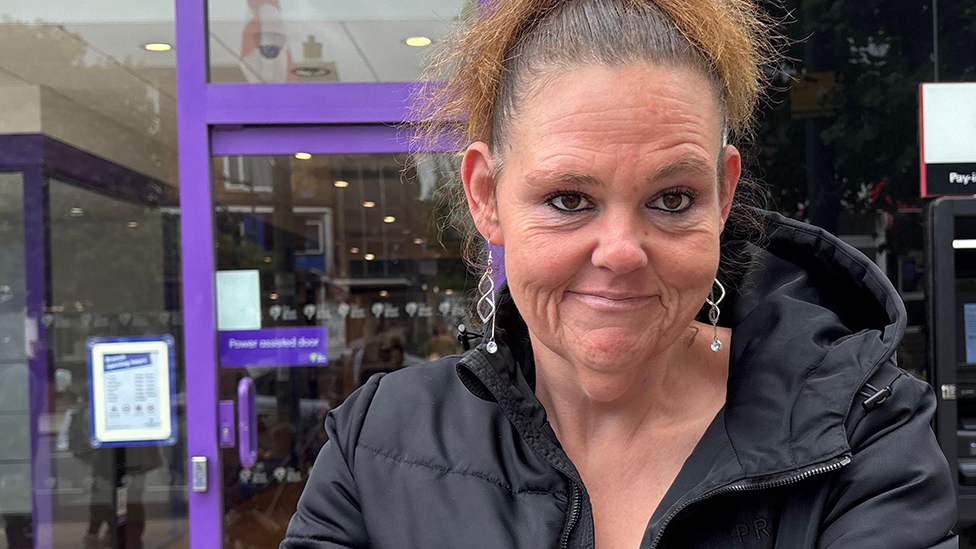 Rebecca Onyeachie is wearing a red top and black puffer jacket. She has a ponytail and is wearing dangly earrings. She has crossed her arms and is smiling at the camera but raising her eyebrows, looking cross. Other high street shops are behind her.