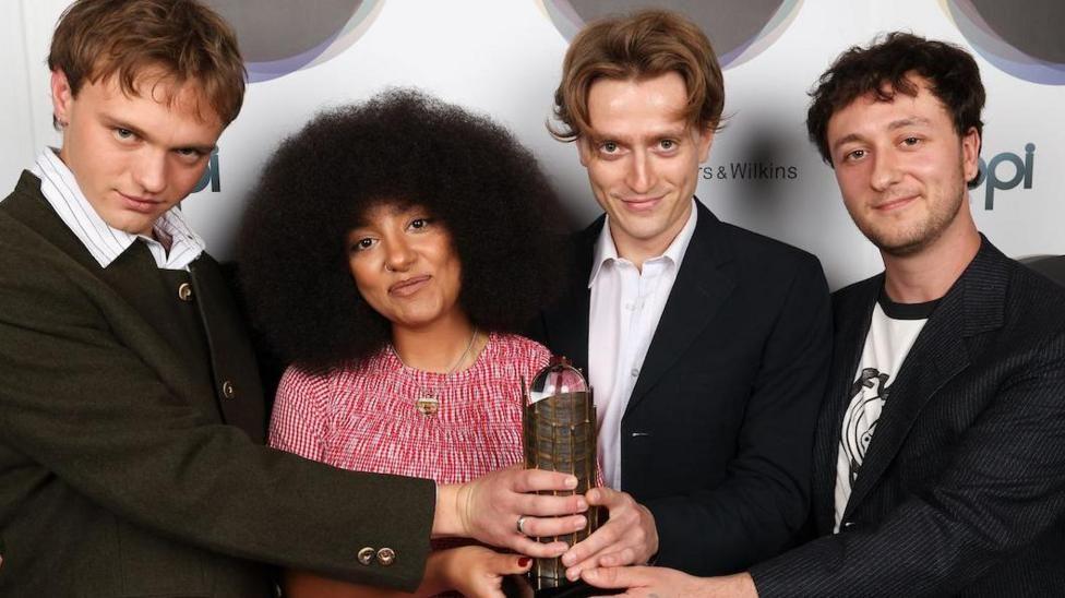 English Teacher with their trophy for the Mercury Prize. The three men in the band are wearing black jackets, two with white button-up shirts and one with a white T-shirt with black design and collar. Second from the left, the female band member has a voluminous black Afro hairstyle and wears a red and white patterned top.