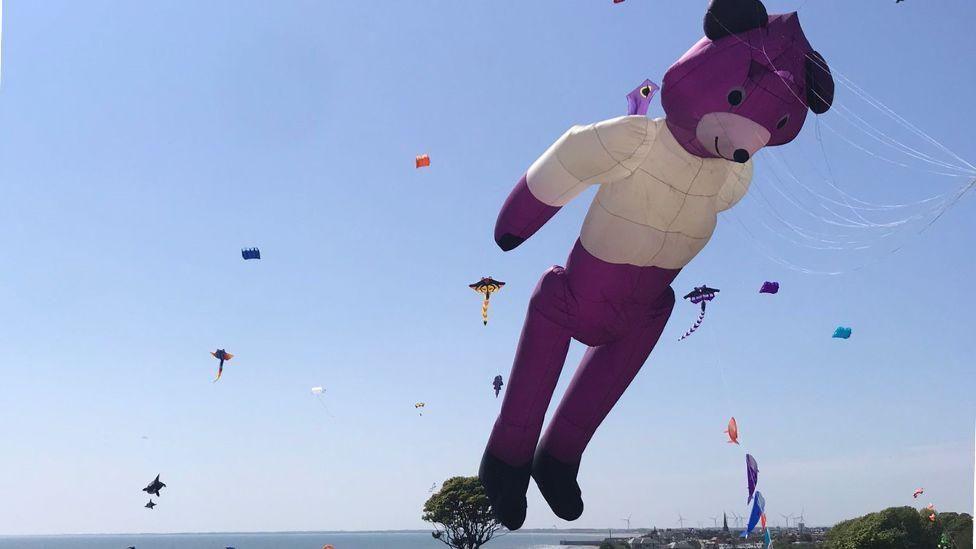 A kite in the shape of a large purple bear flies at the Bridlington Kite Festival. Other colourful kites are visible in the background and are set against a blue, sunny sky.