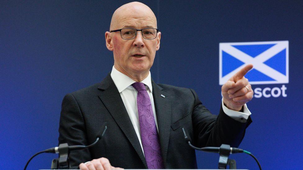 A bald man with glasses points with his left hand while speaking at a podium. He is wearing a dark suit, white shirt and purple tie. There is a blue background behind him with a saltire on it 