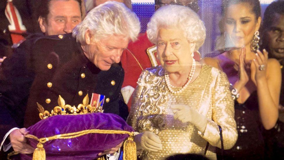 Bruno Peak standing beside Queen Elizabeth at the lighting of her Diamond Jubilee Beacon at the Mall, London. He is on the left and wearing a black jacket with gold buttons and holding a purple gold trimmed cushion with a crown on it. He is leaning towards the Queen, on the left and she is wearing a gold sequinned dress and speaking to him. There are people behind them.