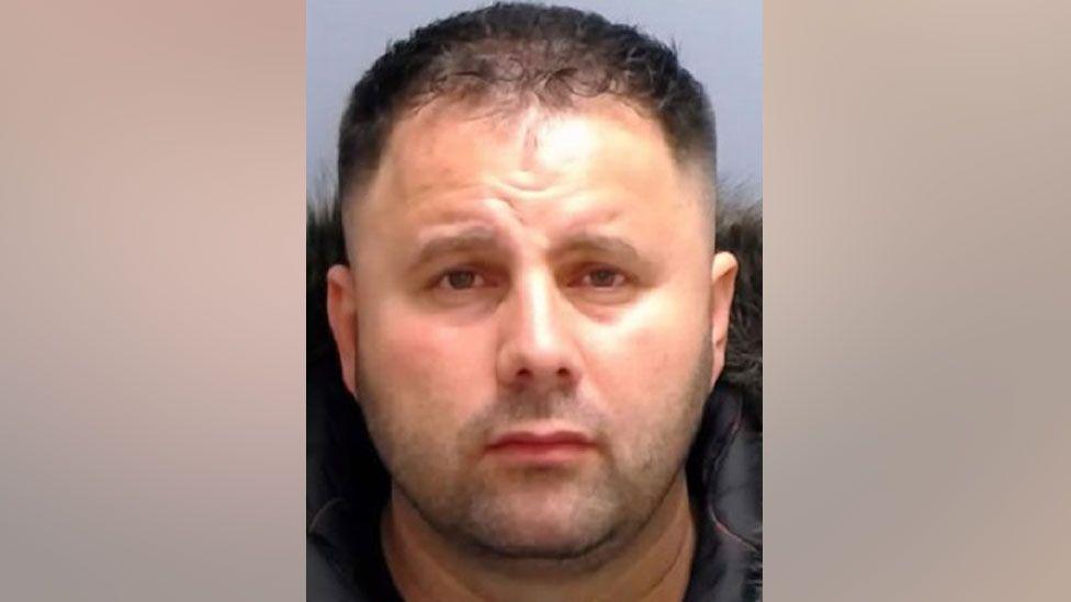 Photo of man looking directly at the camera. He has short black hair, thinning at the front and is not clean-shaven. 
