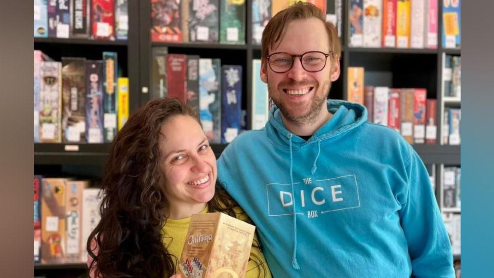 Stephanie and Trevor are standing in front of shelves full of games. Stephanie has brown hair and is wearing a yellow t-shirt, while holding a game named Onitama. Trevor is wearing a blue hoodie and glasses.