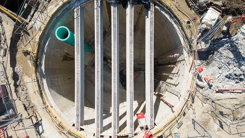 Aerial view of a huge underground storage tank under construction, with five large bars running across a deep round concrete-sided hole.