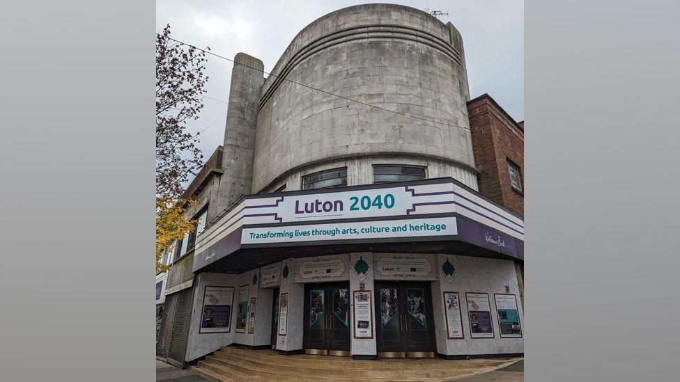 An empty Art Deco cinema in Luton. It shows a building, with light curved brick, with a board with Luton 2040 written on it.  There are windows above and two main doors to the building. There are signs around the front door.