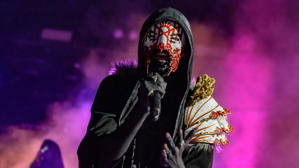 A man singing into a microphone with a black hoodie on, wearing a red white and black mask - with white yellow and red shoulder pads.