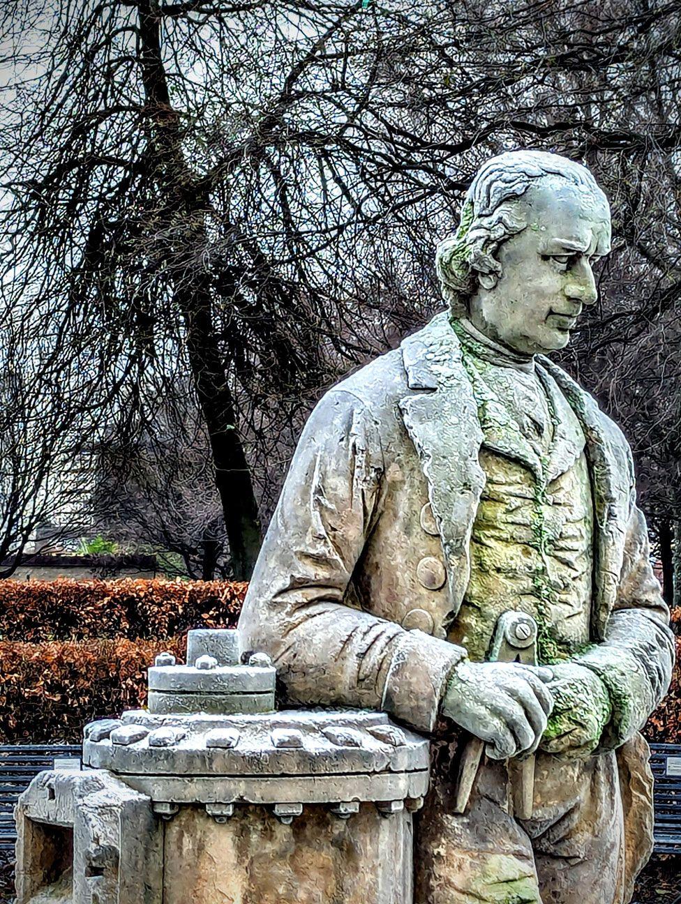 A statue of James Watt looks off into the distance with more than a hint of green on his stone surface due to moss and other coverage. In the background is a very brown-leafed hedge and some bare trees.