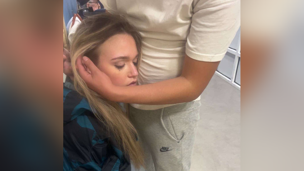 Libby's boyfriend holding her head while she is sat down in the A&E department at the Royal Glamorgan Hospital. She has here eyes closed. Libby has blonde hair and is wearing a blue jacket.