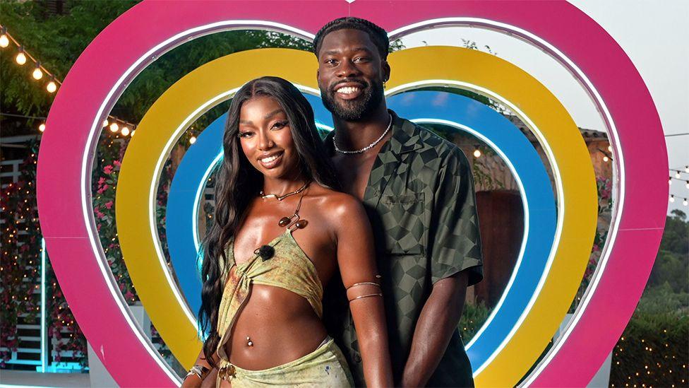 Mimii and Josh, two contestants from Love Island, smiling and standing next to each while a heart-shaped backdrop is behind them in the colours of blue, yellow and pink