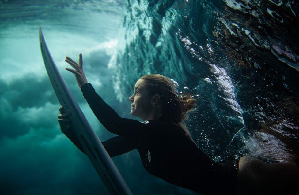 An underwater view of a surfer pushing a surfboard downward while diving beneath a wave. The water is clear with swirling bubbles, and the surfer wears a black wetsuit.