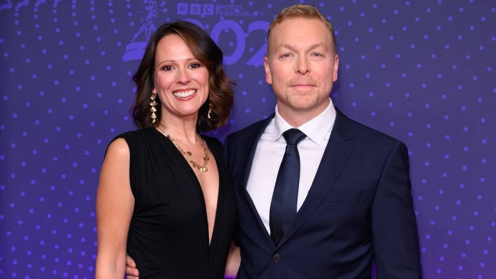 A white woman with short brown hair wearing a black dress and gold jewellery standing with a white man wearing a suit 