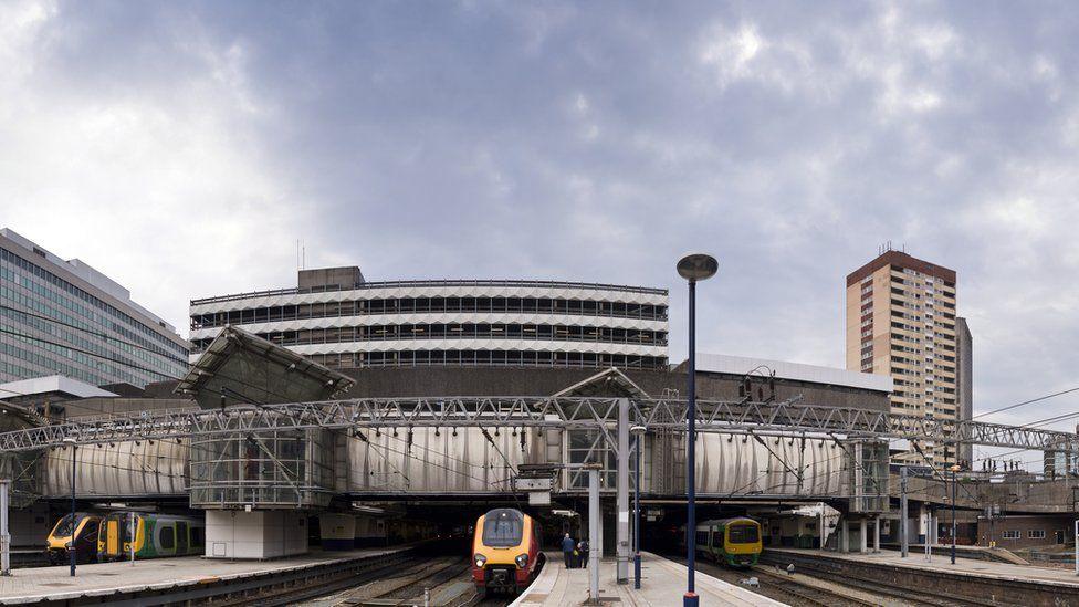 An exterior view of Birmingham New Street station showing multiple tracks and platforms side by side. There are three trains in view on the tracks, with a high rise building to the right in the background
