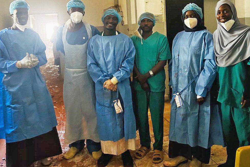 A group of medics at Saudi Hospital in el-Fasher in scrubs smile as they stand for a group photo after successfully performing two-hour emergency caesarean under bombardment. Dust from the shell damage can be seen on the floor