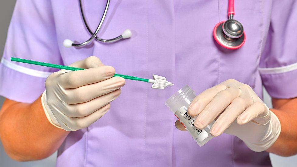 A stock image of a the hands of medical worker in a purple uniform and stethoscope around their neck holding a smear test kit