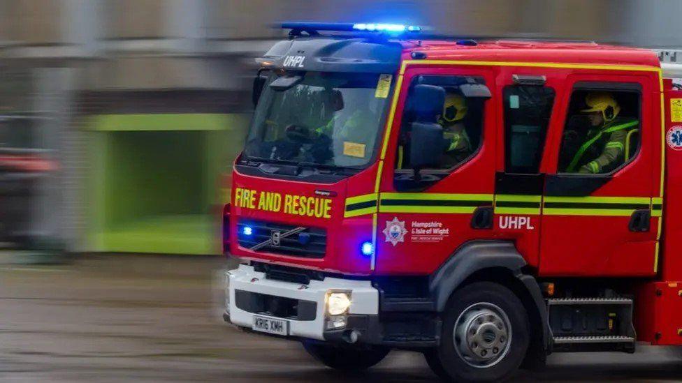 Fire engine pictured travelling from right to left its emergency lights are activated - firefighters can be seen sat in the cab of the appliance.