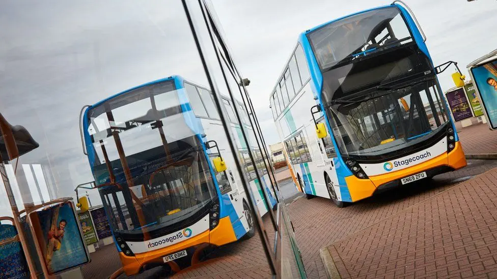An empty blue, white and orange Stagecoach branded double decker bus next to a bus stop. Its reflection can also be seen in the windows of another bus parked next to it, only part of which is in shot.