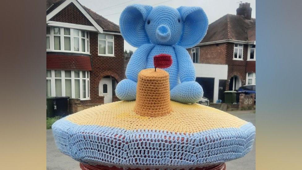 A knitted topper sits on top of a red post box. The topper features blue wool to represent the sea, followed by beige wool, depicting sand. On the sand, is a small knitted sandcastle with a red flag, and behind it is a blue knitted elephant.