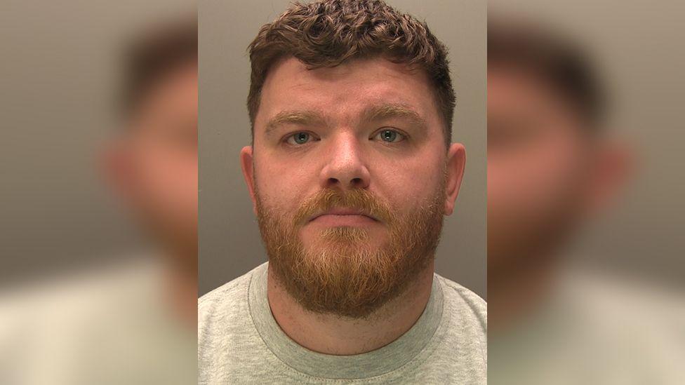 Police custody photograph of Stuart Wells who has dark short hair and beard and is wearing a grey shirt