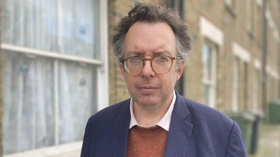 Richard Blakeway is pictured outdoors in front of a row of brick terraced houses. He is wearing glasses, a navy jacket and an orange jumper, looking directly at the camera.