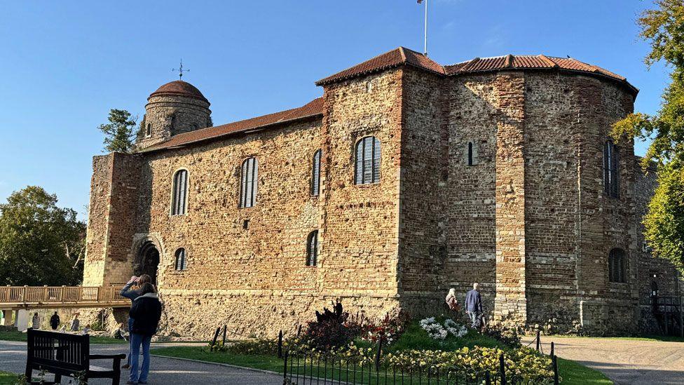 Colchester Castle, a golden coloured castle, originally Norman built, with small glazed windows on its ground floor and large glazed windows on its first floor. There is a squat round tower at its left side and the roof is of the building is tiled in brown