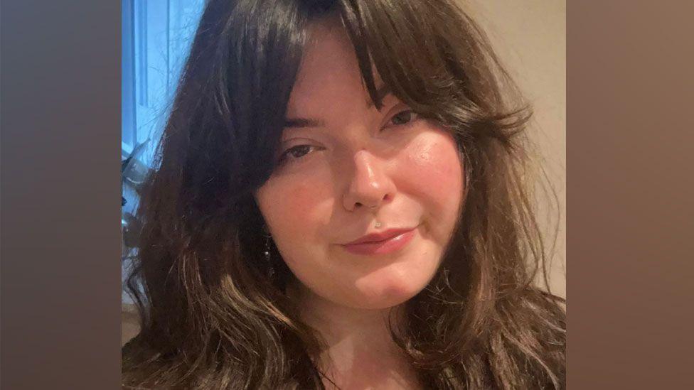 Chloe Humphries who has long dark wavy hair over her shoulders. Her head is tilted to her right and she has a serious expression on her face.