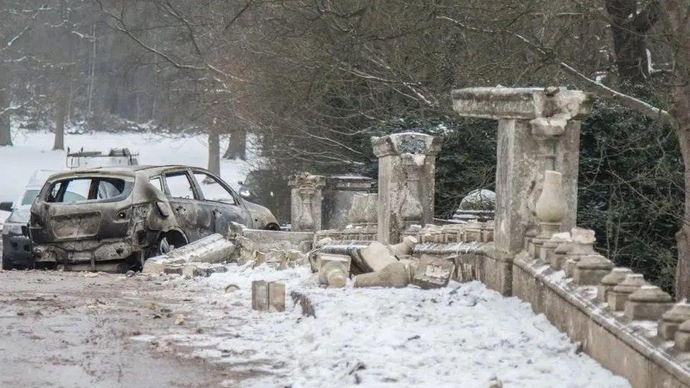 A burnt-out car sat next to ruined stonework of a bridge in heavy snow