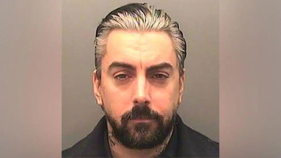 A mugshot of Ian Watkins - a man with greying slicked back hair and a shortish dark beard. He is staring straight into the camera.