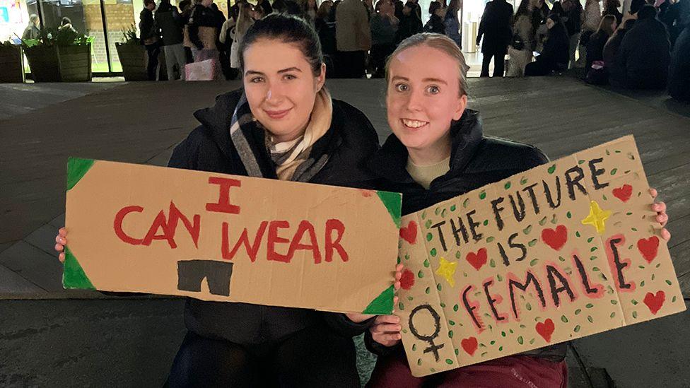 Two women dressed in dark coats hold handmade cardboard signs one saying "I can wear shorts" the other saying "The future is female"