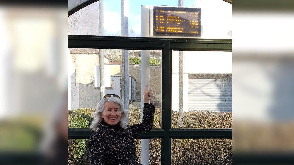 Wendy is standing in a bus shelter near the window. It is a sunny day outside. She is smiling and pointing up to a bus sign with words that are unreadable. She wears a black jacket with a colourful diamond pattern on it and black sunglasses perched on her head. She has white hair. 