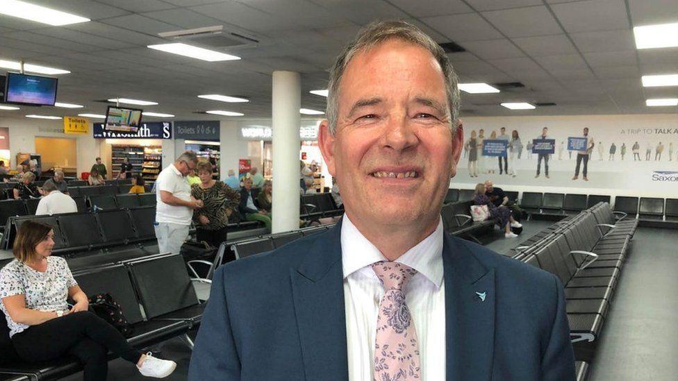 Richard Pace wearing a blue suit jacket, white shirt and pink and grey floral tie stands inside Norwich Airport lounge. There are passengers in the background, some are seated.