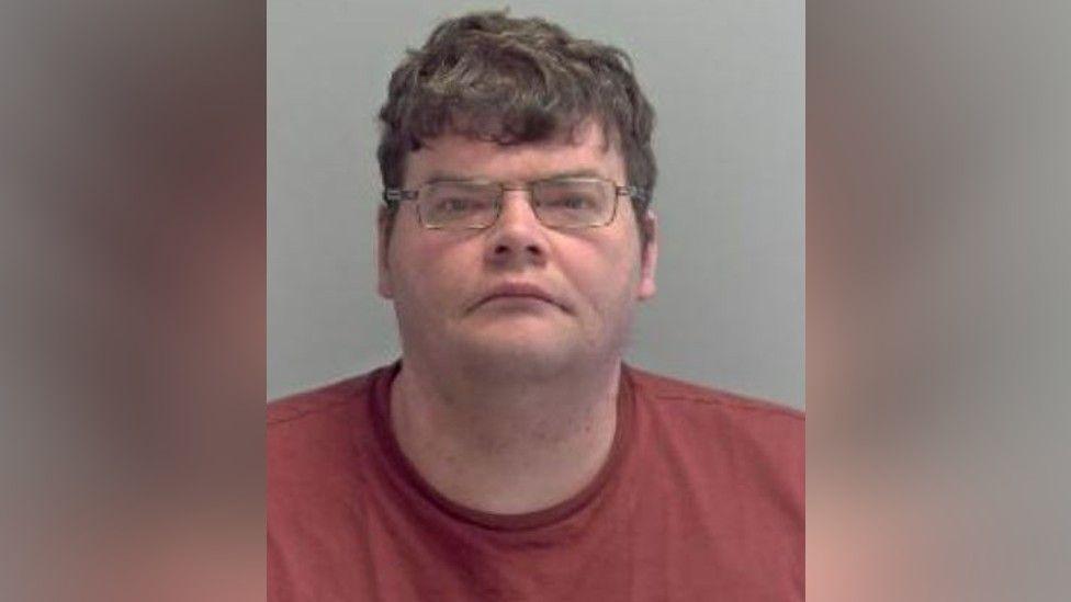 Police custody photograph of Daniel Russell. He is wearing glasses and is  looking at the camera. He is wearing a red t-shirt.