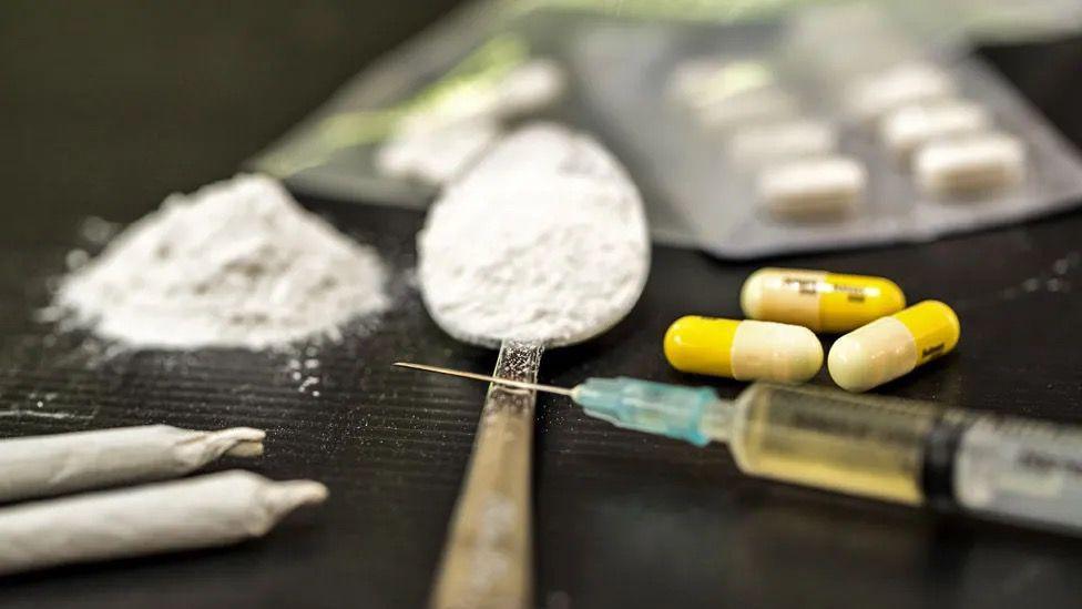 Drugs are laid out on a black surface, with powder on a spoon, yellow pills, a liquid in a syringe