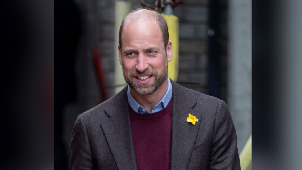 Prince William wearing a black jacket with a daffodil on the left lapel, he has a maroon jumper underneath a blue shirt under that 