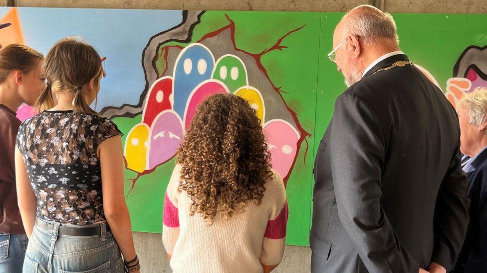 The back view of three teenagers and a man wearing a mayoral chain over his grey suit and a woman with grey hair looking at part of the finished mural 