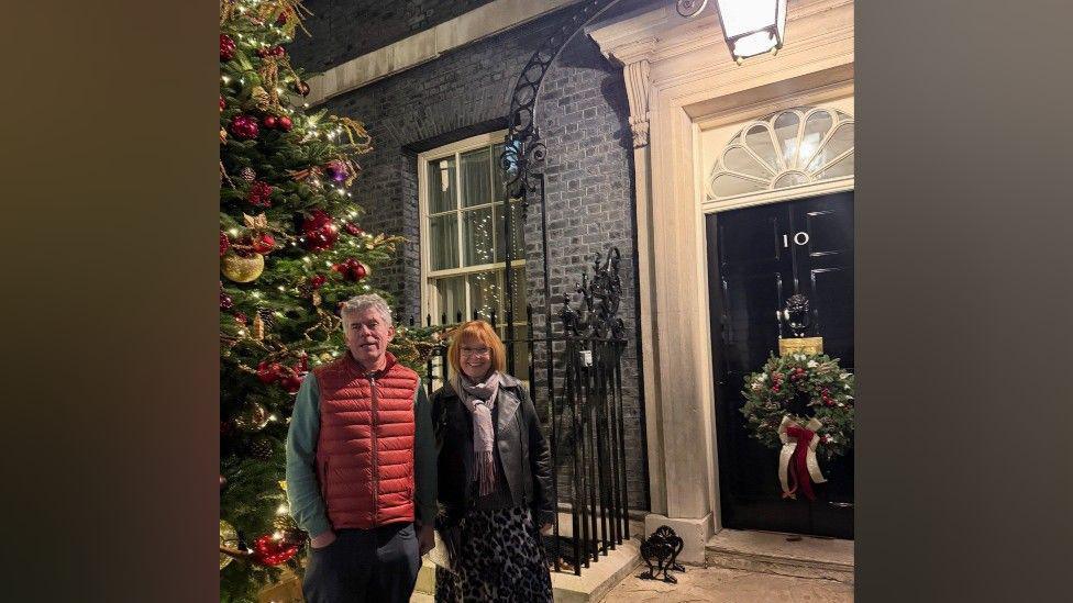 A man and a woman are standing in front of the door to 10 Downing Street. There is a large fir Christmas tree to their left, covered in decorations. There is a large wreath on the door of the building, with a red and white ribbon trailing down from it