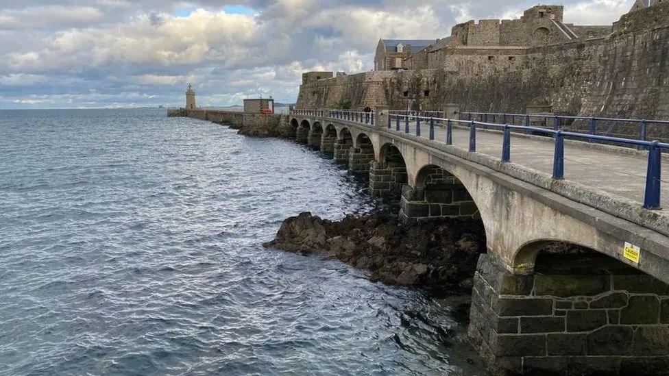 A long multi-arched bridge which leads to a lighthouse and a castle with the sea to its left.