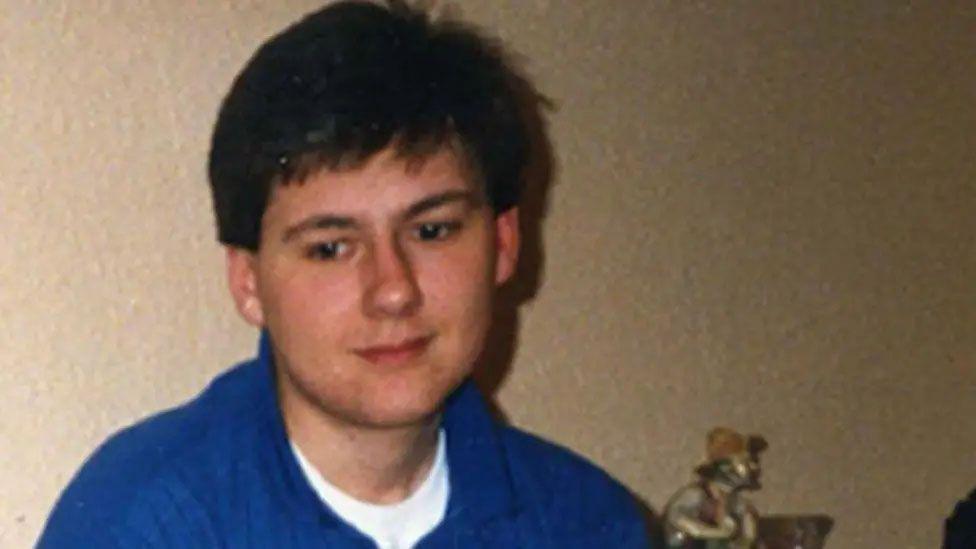Christopher has short dark hair and is wearing a blue jumper over a white t-shirt.