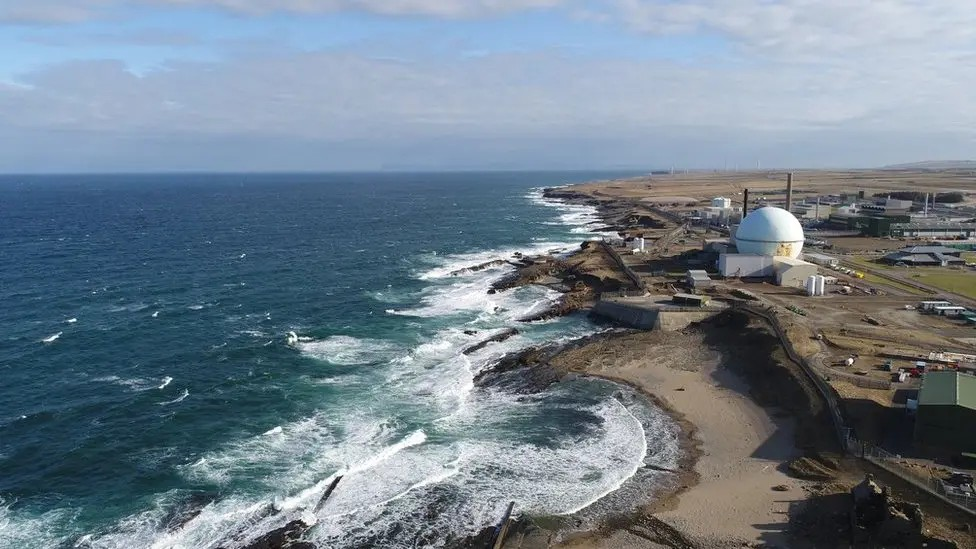 The power station is a complex of buildings and structures, including metal sheds with peaked roofs, a chimney and a large sphere. It is set by the sea, with a sandy shore pictured between white waves and nuclear facility.