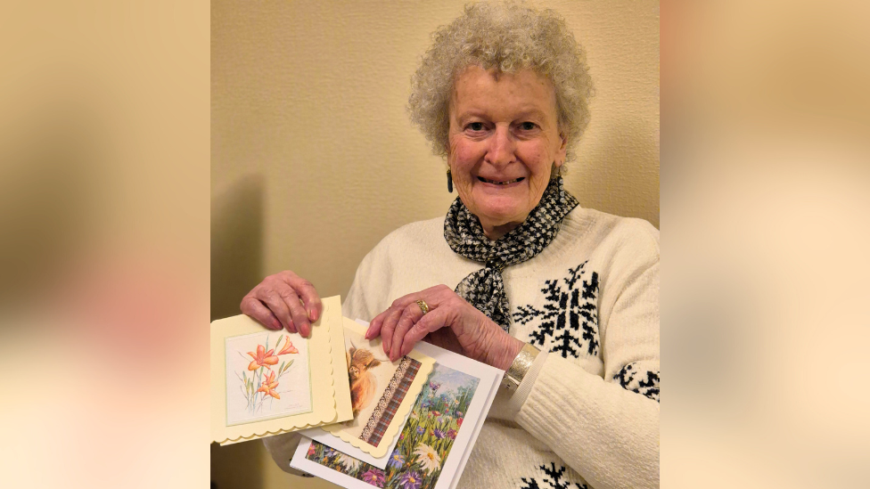 An older woman, wearing a white jumper, holds up colourful homemade cards and smiles.