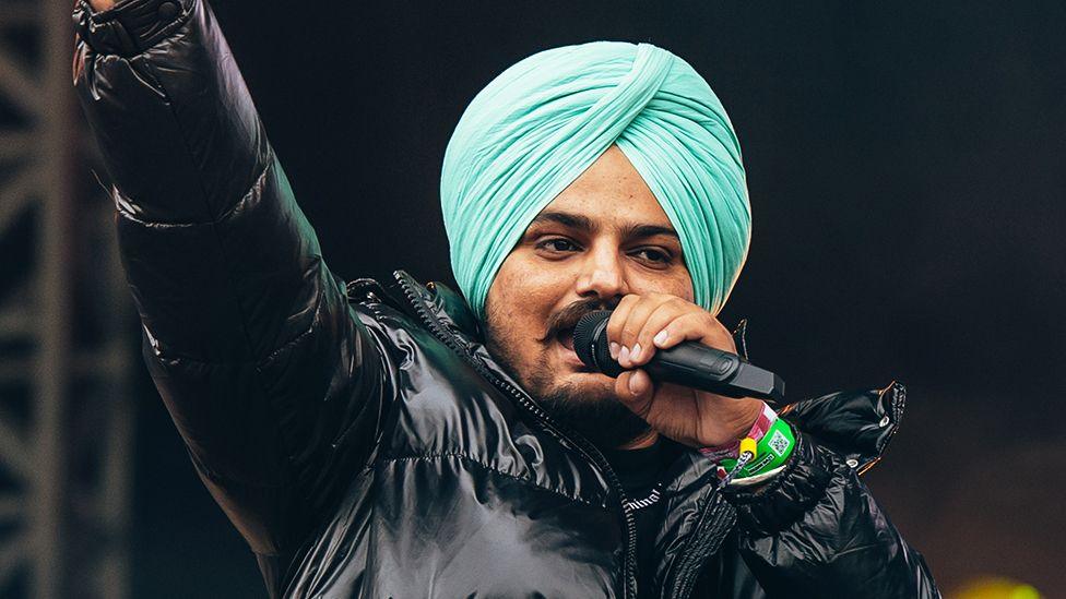 A man wearing a black jacket, performing on stage singing into a black microphone, wearing a light blue turban (religious head covering for Sikh's).