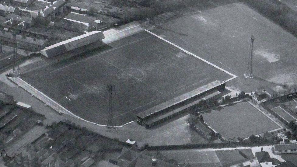 A black and white aerial shot of Cambridge City's Milton Road ground in 1960. The ground takes up much of the shot and shows two covered stands at its top and bottom sides. Tall scaffolding towers for spot lights can be seen at all corners of the ground. On the top left and along the bottom can be seen houses and gardens 