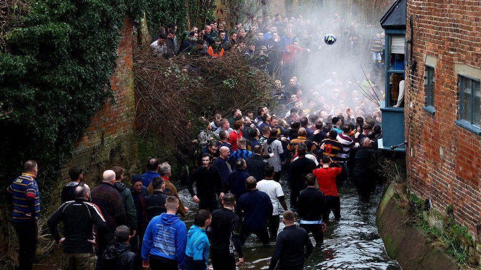 Huge crowds stand in a brook between buildings during the game, a ball can be seen in the air above the players